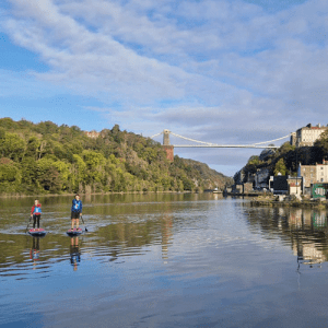 two paddleboarders paddle under the Clifton Suspension Bridge in Bristol on a sunny calm day at high tide.