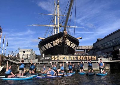 A large paddleboarding group of men and women outside Brunels' SS Great Britain ship on Bristol Harbour.