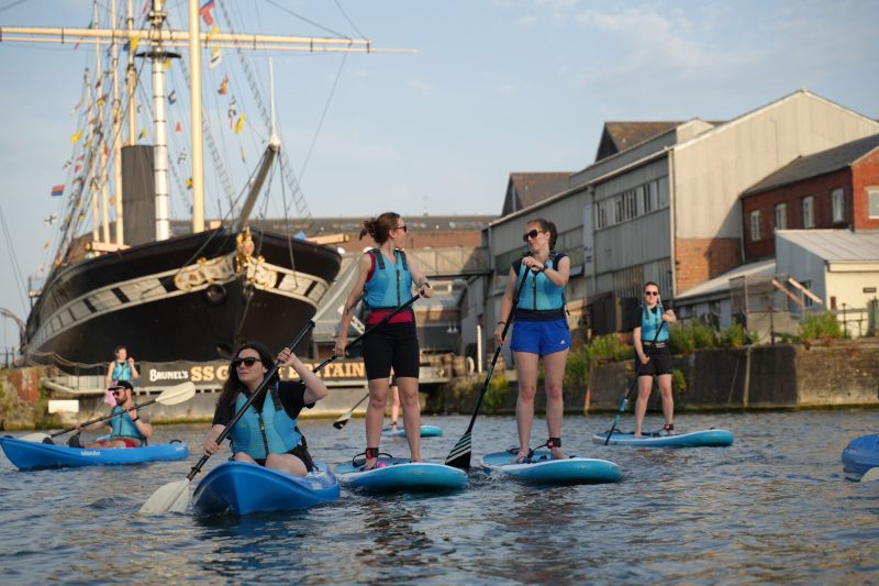 A mixed group of people are on the water of Bristol Harbour; the SS Great Britain tall ship is docked behind the group. Everyone is wearing buoyancy aids, and some are sitting on sit-on-top kayaks and others are standing up on paddleboards as they paddle around the harbour.