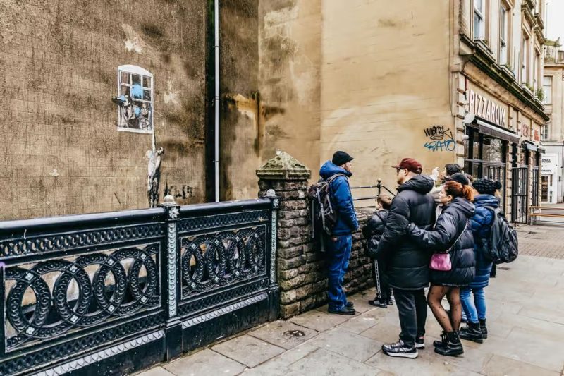 A group of people on a tour stand in front of a stone wall, looking at Banksy’s ‘Well-hung Lover’ graffiti piece on Frogmore Street in Bristol.