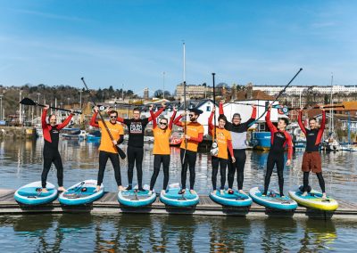 A mixed group of people stand in a row on their paddleboards, hoisting their paddles overhead as they smile and cheer. They are on a narrow dock over the water on Bristol Harbour, with boats moored behind them. They are all wearing wetsuits, and some are wearing orange t-shirts over their wetsuits.