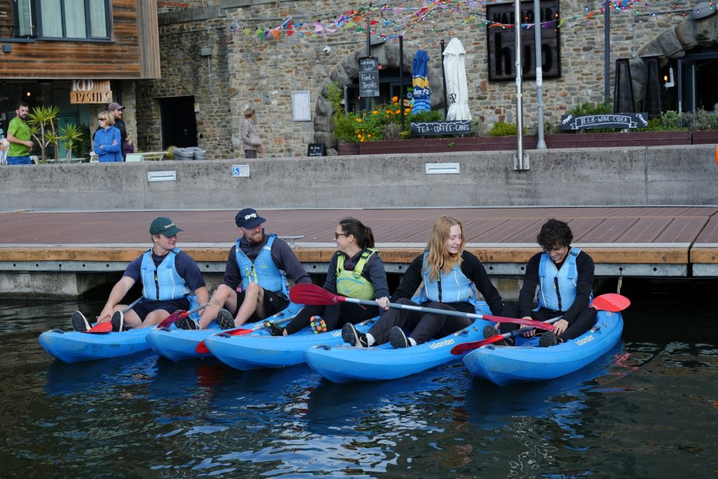 A mixed group of men and women settle into their sit-on-top kayaks at a dockside in Bristol. They are wearing buoyancy aids and holding kayak paddles as they smile and chat with each other before setting off.