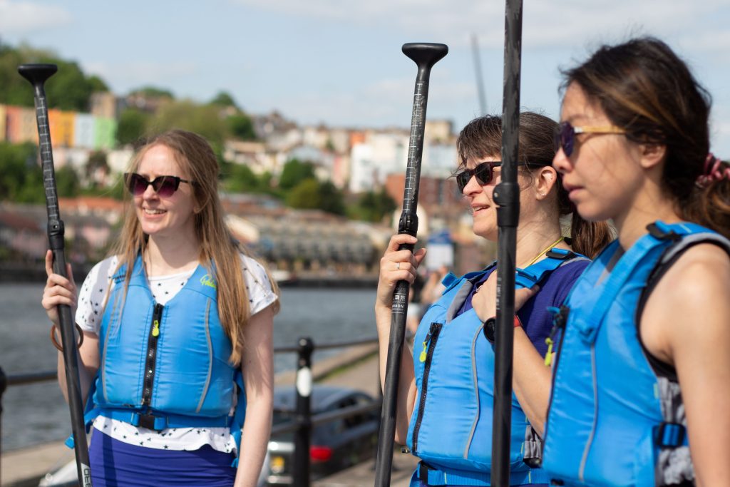 Three women stand in a row, listening to the pre-session brief. They are wearing buoyancy aids and holding their SUP paddles. All three are wearing sunglasses and smiling. They have joined a public group session paddling Bristol Harbour.