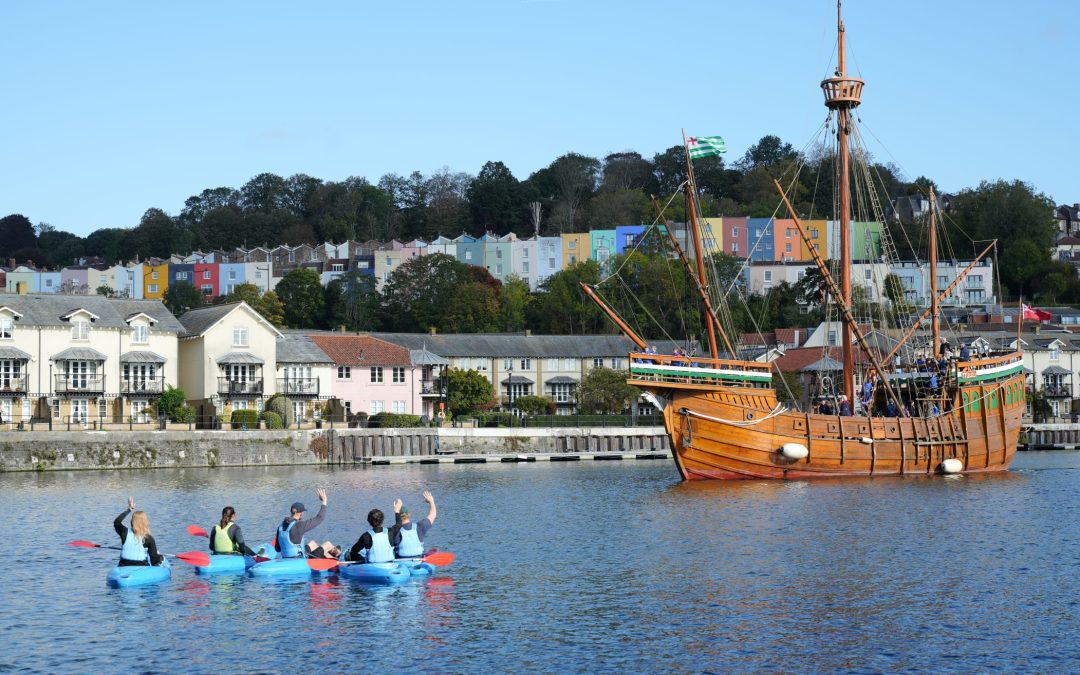 A mixed group of people are trying a hen or stag group kayaking activity on Bristol Harbour. They are waving at a historic replica of a tall ship as it makes its way through the harbour.