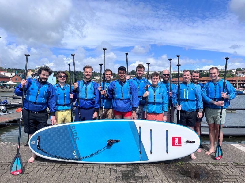 A large group of men are smiling, holding SUP paddles and standing on the wharf at Bristol Harbour, about to embark on a stag party paddleboarding activity with SUP Bristol.