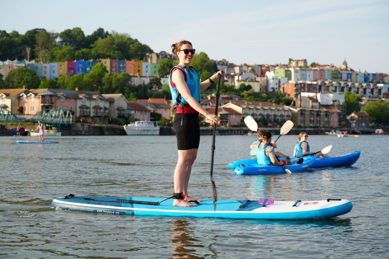 A woman stands on a paddleboard in Bristol Harbour and smiles. She is wearing a blue buoyancy aid, holding a black paddle, and has the board's safety leash around her ankle. In the background are three people on sit-on-top kayaks, and the colourful terrace houses of Bristol rise up in the background.