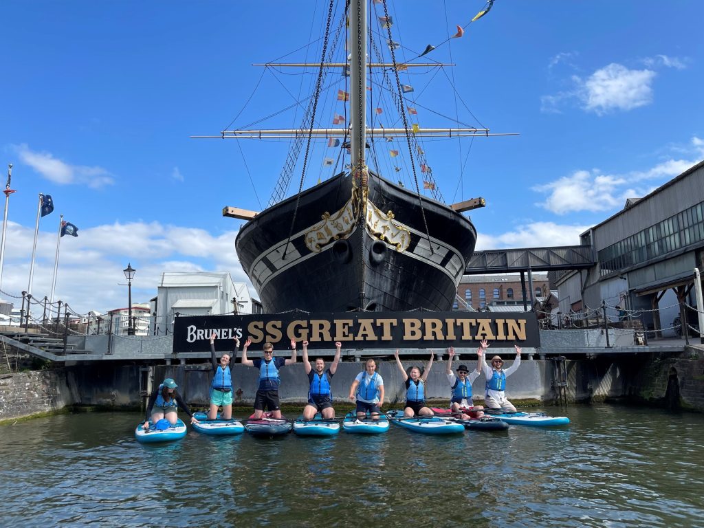 A mixed group of 8 people are lined up on the water on their paddleboards in front of the famous SS Great Britain, a historic ship and popular sightseeing destination, designed by Isambard Kingdom Brunel and moored in Bristol Harbour.