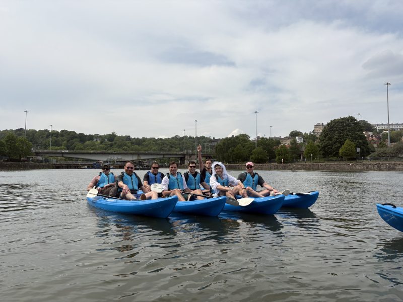 A group of men on a stag do sit on kayaks on the water at Bristol Harbour smile at the camera. One is dressed in a wedding dress costume, getting ferried around by his mates.
