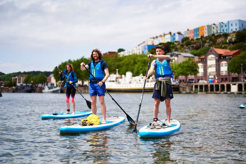 Three adults smile as they paddle through Bristol Harbour on stand-up paddleboards. Historic buildings line the harbourside, and the colourful terrace houses of Bristol stand proudly on the horizon.