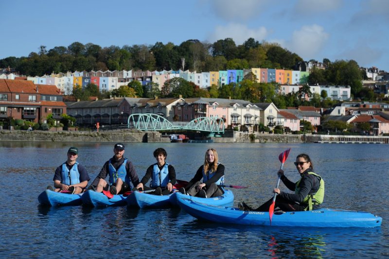 Five adults on sit-on-top kayaks are lined up on the water of Bristol Harbour for a photo. They are smiling, and you can see the historic harbour-side buildings in the background.